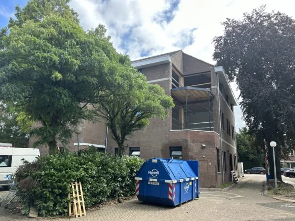 Bakstenen kantoorgebouw aan de Bergse Dorpsstraat met een blauwe afvalcontainer en omliggende bomen.